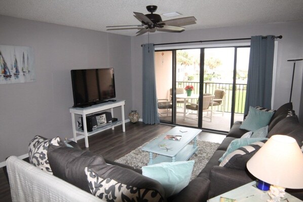 A living room with a sofa, coffee table, TV, and a balcony with outdoor furniture. - Sea Place 13137 2 Bedroom Condo (St. Augustine)
