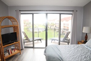 A hotel room with a bed, a television on a stand, a balcony with chairs, and a view of the surrounding buildings. - Sea Place 13137 2 Bedroom Condo (St. Augustine)