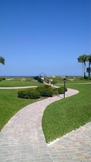 A paved pathway curving through a well-maintained grassy area with a clear view of the ocean in the background. - Sea Place 13137 2 Bedroom Condo (St. Augustine)