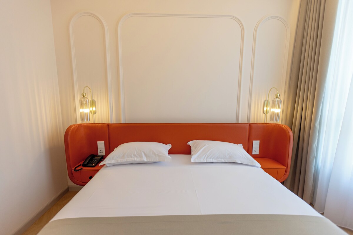 Quarto de hotel com uma cama grande, duas mesinhas de cabeceira, telefone e iluminação embutida na parede.