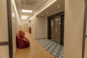 Corredor de hotel com design moderno, piso estampado, área de estar com cadeiras vermelhas e um elevador.