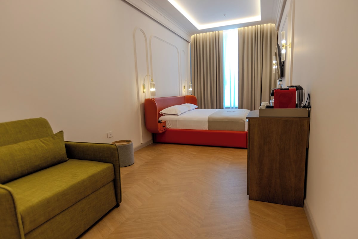 Quarto de hotel com cama, sofá, escrivaninha e máquina de café, além de um espelho.