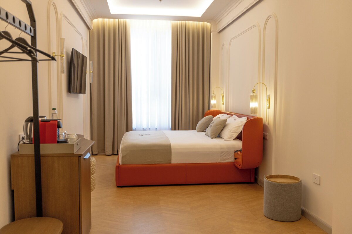 Quarto de hotel com cama, escrivaninha, cadeira, mesa pequena, lixeira e televisão na parede.