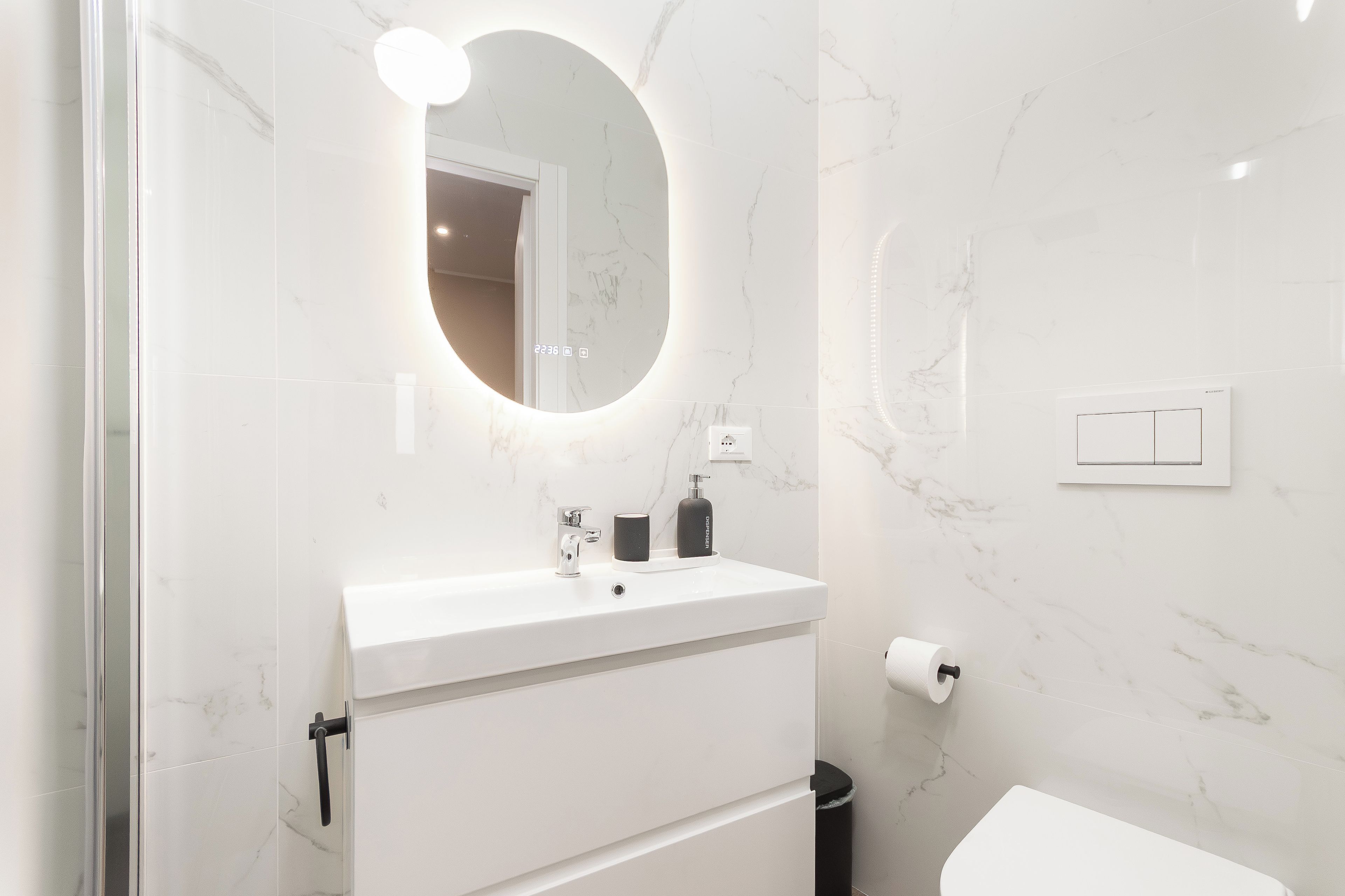 Ein modernes Badezimmer mit einem weißen Marmorwaschbecken, einem runden Spiegel, einem schwarzen Seifenspender und einer Toilettenpapierrolle.