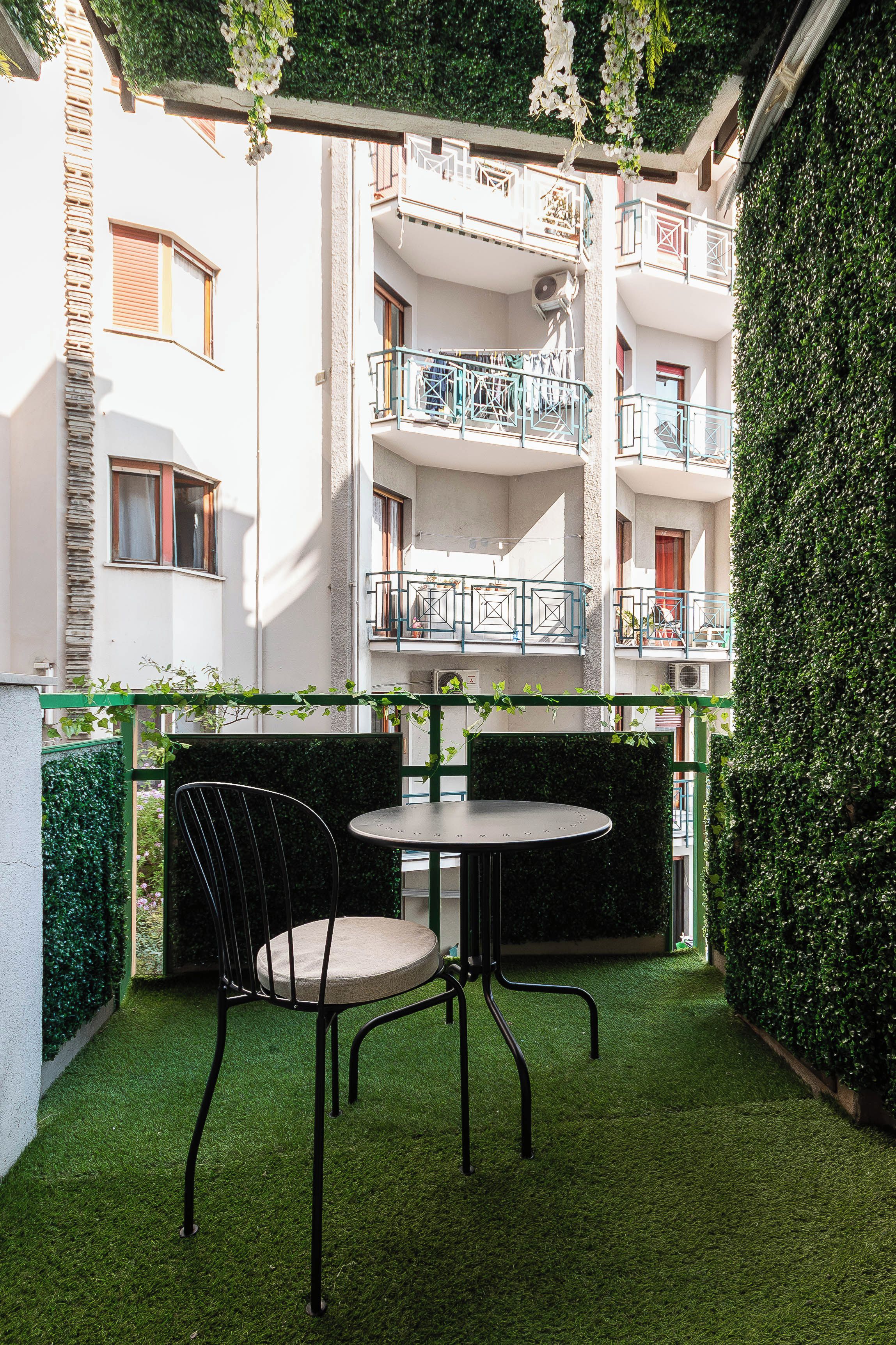 A courtyard with a table and chairs, surrounded by a hedge and a building with balconies.