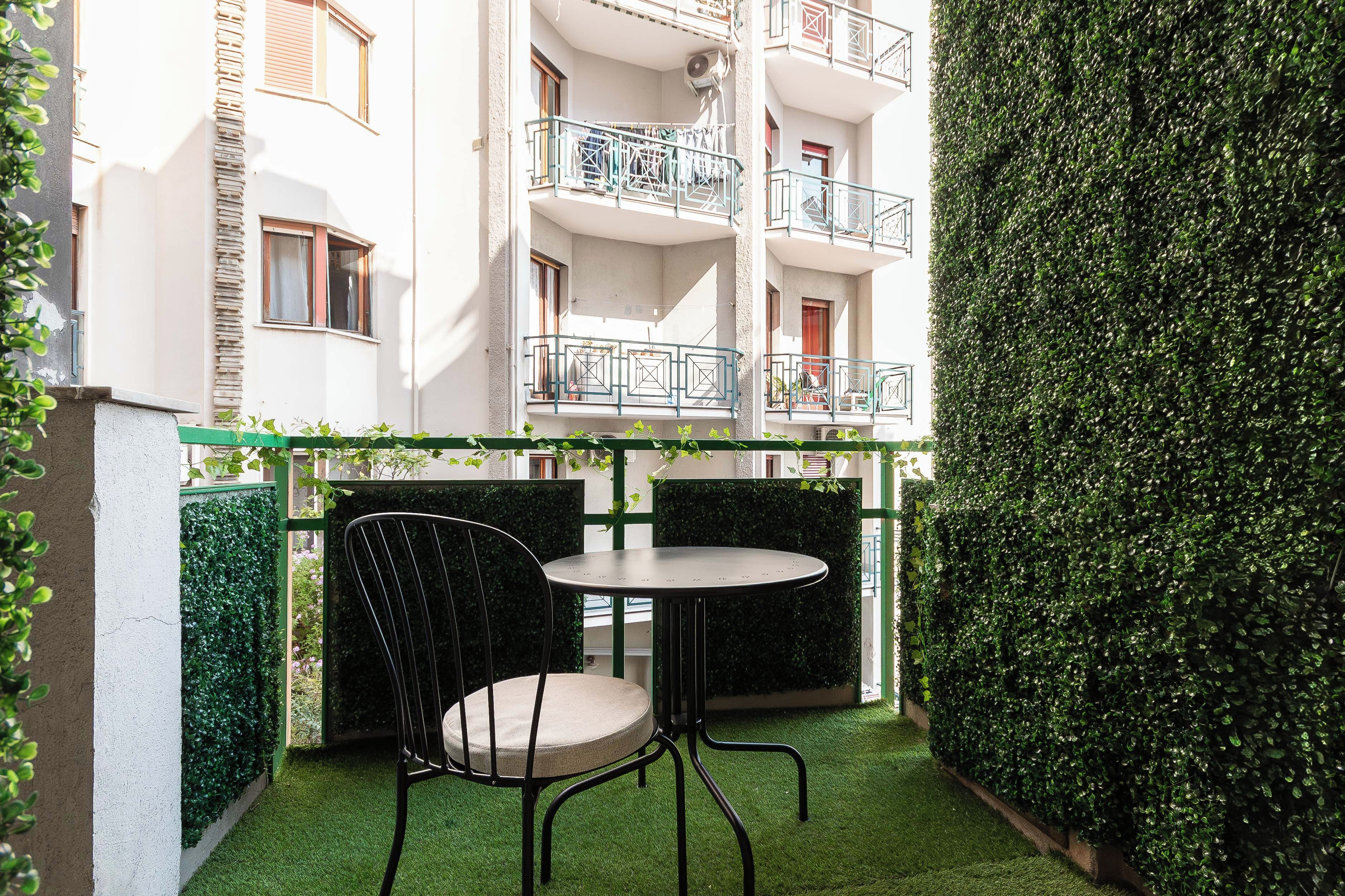 Un petit espace extérieur aménagé avec une table et deux chaises, entouré d’une haie verdoyante et d’un mur blanc.