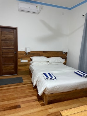 A hotel room with a wooden floor, a bed with white bedding, a blue-striped towel, a wooden door, a wall-mounted air conditioner, and a power outlet. - Mandhoo Inn (Mandhoo)