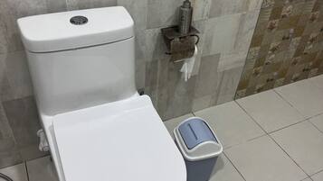 A white toilet with a flush handle, a small trash bin, and a paper towel dispenser mounted on the wall.
