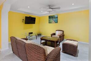 A living room with a flat-screen TV mounted on the wall, a glass coffee table, wicker furniture, and a ceiling fan. - Hospitality Expert VSV BF 3BR Chef Pool  (Montego Bay)