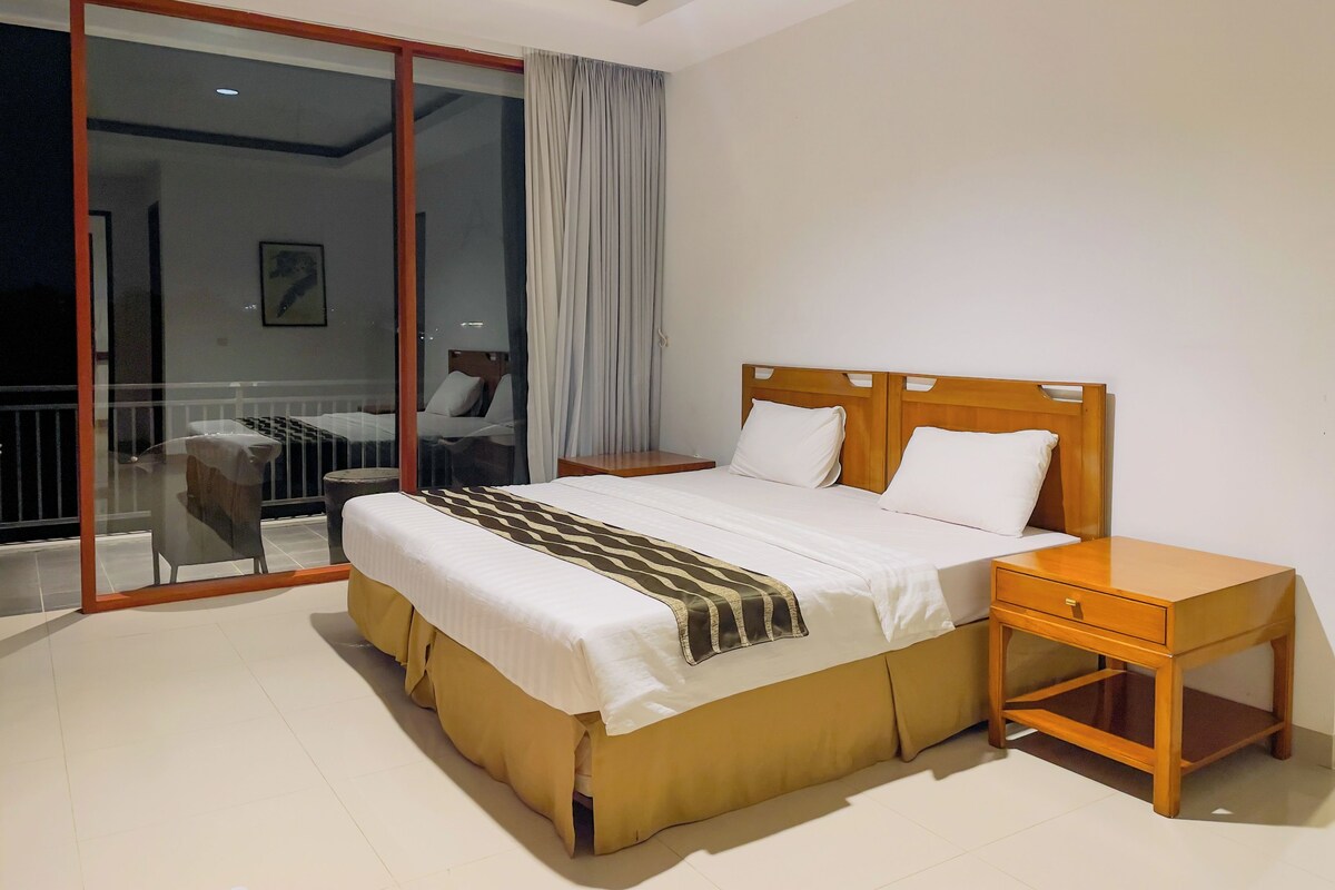A hotel room with a bed, bedside table, and a view through a glass door to an outdoor area.