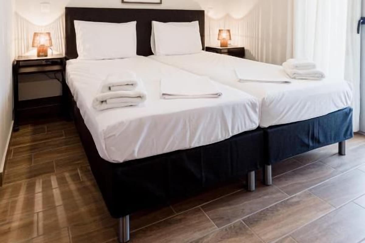 Una cama doble con cabecera negra, dos mesitas de noche con lámparas, un ventilador de techo y un cuadro enmarcado en la pared.