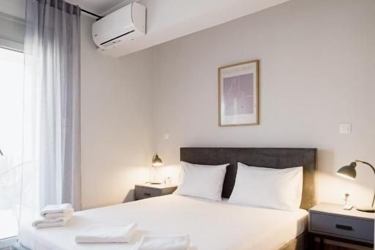 Una habitación de hotel con una cama, un ventilador de techo, un equipo de aire acondicionado, una mesita de noche con una lámpara y un cuadro en la pared.