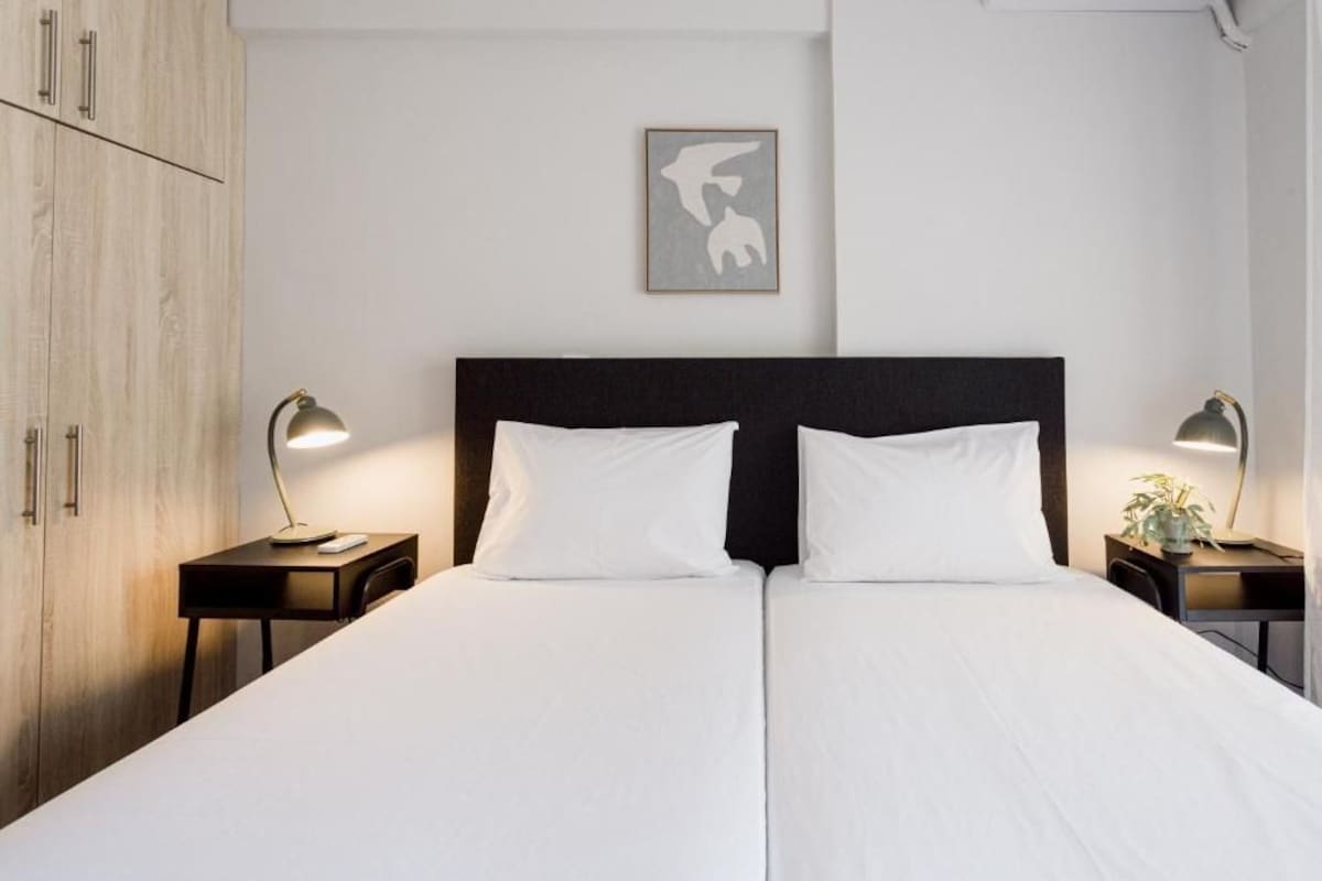 Una cama doble con ropa de cama blanca, dos mesitas de noche con lámparas, un armario de madera y una obra de arte enmarcada en la pared.