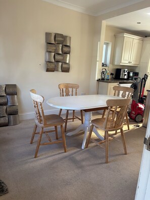 A dining area with a round table and four wooden chairs, a kitchen in the background, and a decorative wall piece.