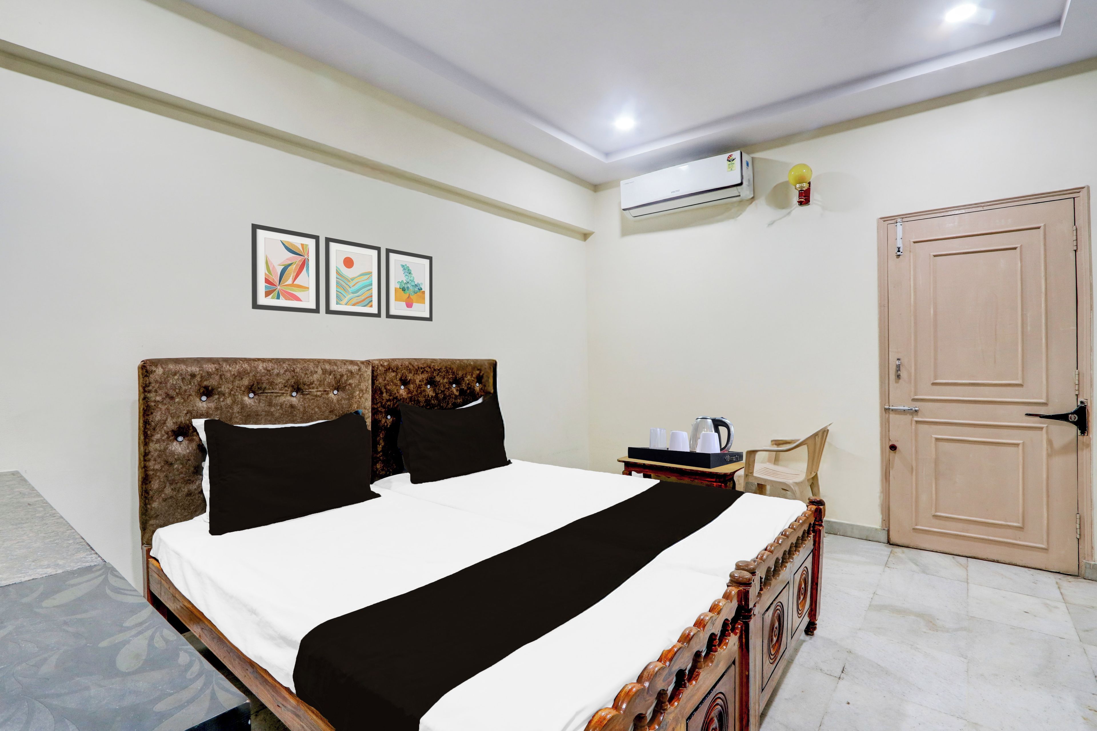A neatly made bed with a headboard, a bedside table with a kettle and cups, and a door to the right.