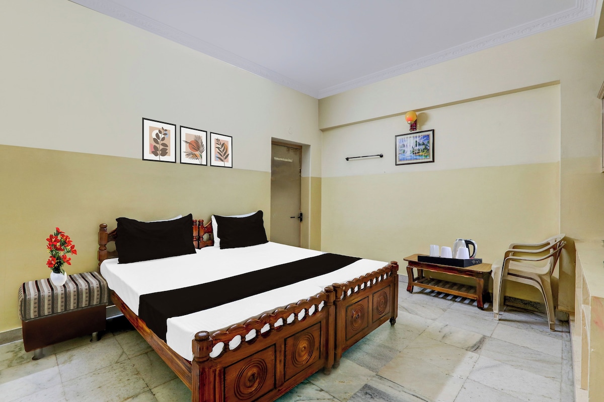Une chambre d’hôtel avec un lit en bois, une table de chevet avec une lampe, une petite table avec des tasses et une bouilloire, et une chaise.