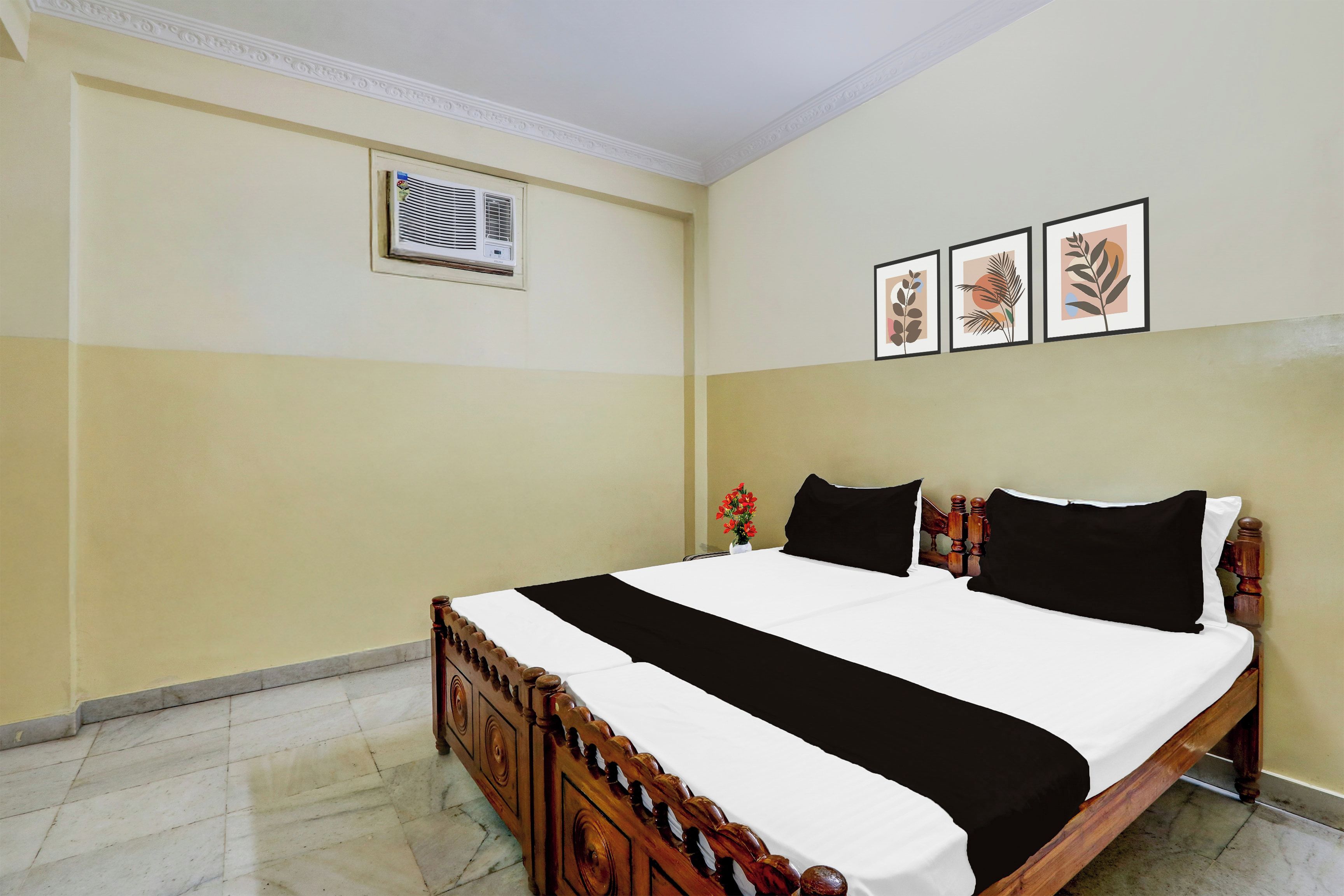 A hotel room with a bed, a wall-mounted air conditioning unit, and three framed botanical prints.