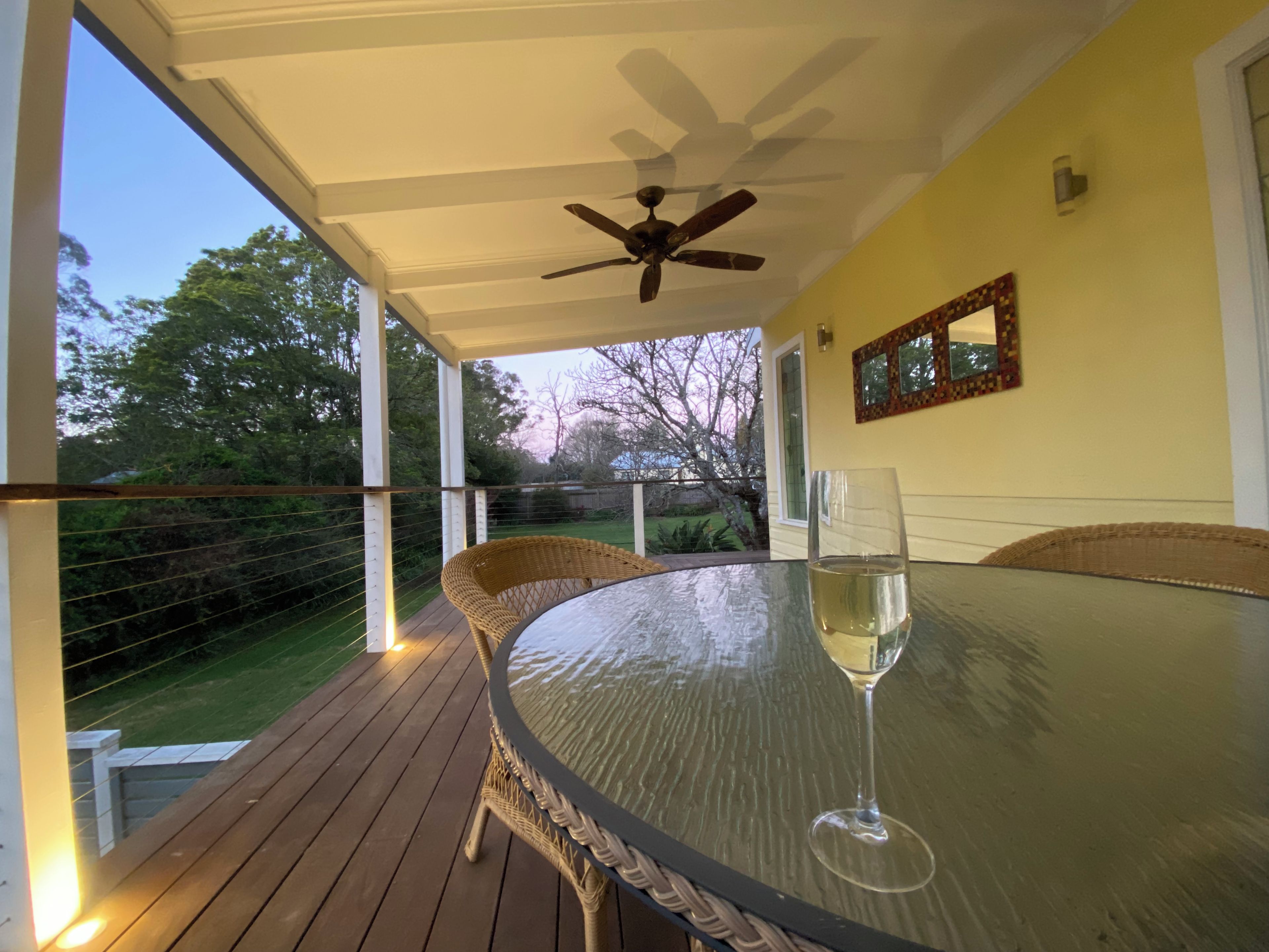 A covered outdoor patio with a glass table, wicker chairs, a ceiling fan, and a glass of wine.