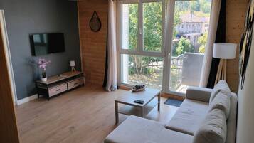 Een moderne woonkamer met een flatscreen televisie, houten vloer, een bank, een salontafel en een groot raam met uitzicht op groen en gebouwen.