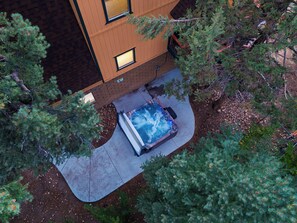 A hot tub in a landscaped area with a wooden building and surrounding trees. - Hot Tub + Firepit + Grill | Mountain Views + Mins to Slopes | Bear Pine Lodge (Big Bear)