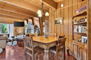 A cozy lodge interior with a wooden dining table, chairs, a fireplace, a bookshelf, and a television mounted on the wall. - Hot Tub + Firepit + Grill | Mountain Views + Mins to Slopes | Bear Pine Lodge (Big Bear)