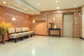 Uma área de recepção com parede de tijolos, um banco, um vaso com flores, uma mesa pequena e um elevador.