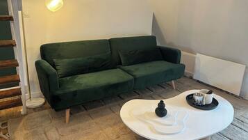 Ein grünes Sofa, ein weißer Couchtisch mit einem schwarzen Gegenstand und einer Tasse, sowie eine Stehlampe in einem Raum mit geneigter Decke.