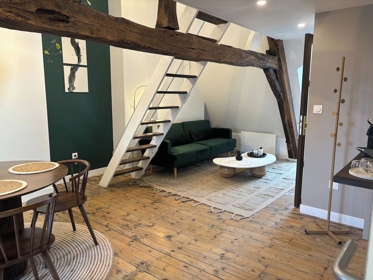 Een loft-achtige woonruimte met zichtbare houten balken, een groene bank, een eettafel met stoelen en een witte salontafel.