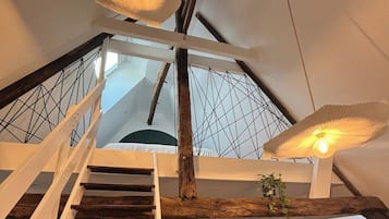 Un intérieur à plusieurs niveaux, doté d’un escalier en bois, d’un coin repas avec une table ronde et offrant une vue sur le ciel grâce à un motif géométrique ornant le plafond.