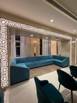A modern living room with a blue sectional sofa, two wall-mounted light fixtures, and a decorative geometric pattern on the walls. - Hotel Samrat Sheraton (Varanasi)