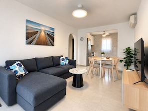 Una sala de estar moderna con un sofá seccional, una mesa de centro redonda, una zona de comedor con mesa y sillas, un televisor y un cuadro en la pared.