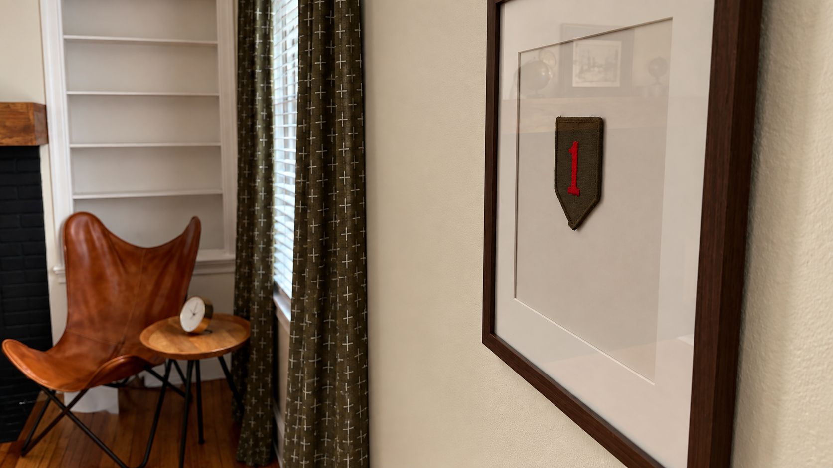 Un parche militar enmarcado en una pared, un sillón de cuero marrón, una mesita redonda con un reloj y una ventana con cortinas.