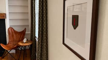 Un parche militar enmarcado en una pared, un sillón de cuero marrón, una mesita redonda con un reloj y una ventana con cortinas.
