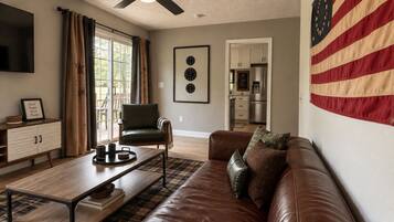 A living room with a brown leather sofa, a wooden coffee table, a green leather chair, and a kitchen in the background.