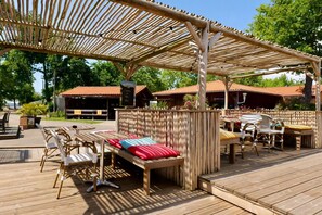 An outdoor seating area with wooden tables and chairs, a covered section with a pergola, and a building with a red roof in the background. - PREMIUM 6-PERSON MOBILE HOME WITH 3 BEDROOMS AT 5-STAR MAYOTTE CAMPSITE—AIR CONDITIONING, FURNISHED, PARKING, LAKE ACCESS (Biscarrosse)