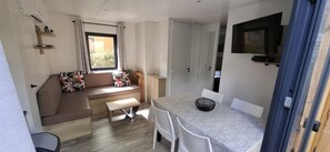 A compact living space with a sofa, a dining table, and a television. - PREMIUM 6-PERSON MOBILE HOME WITH 3 BEDROOMS AT 5-STAR MAYOTTE CAMPSITE—AIR CONDITIONING, FURNISHED, PARKING, LAKE ACCESS (Biscarrosse)