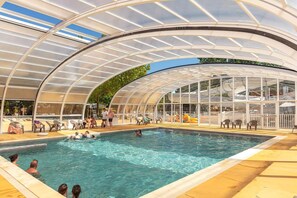 An indoor swimming pool with a glass ceiling, surrounded by lounge chairs and a poolside area. - PREMIUM 6-PERSON MOBILE HOME WITH 3 BEDROOMS AT 5-STAR MAYOTTE CAMPSITE—AIR CONDITIONING, FURNISHED, PARKING, LAKE ACCESS (Biscarrosse)