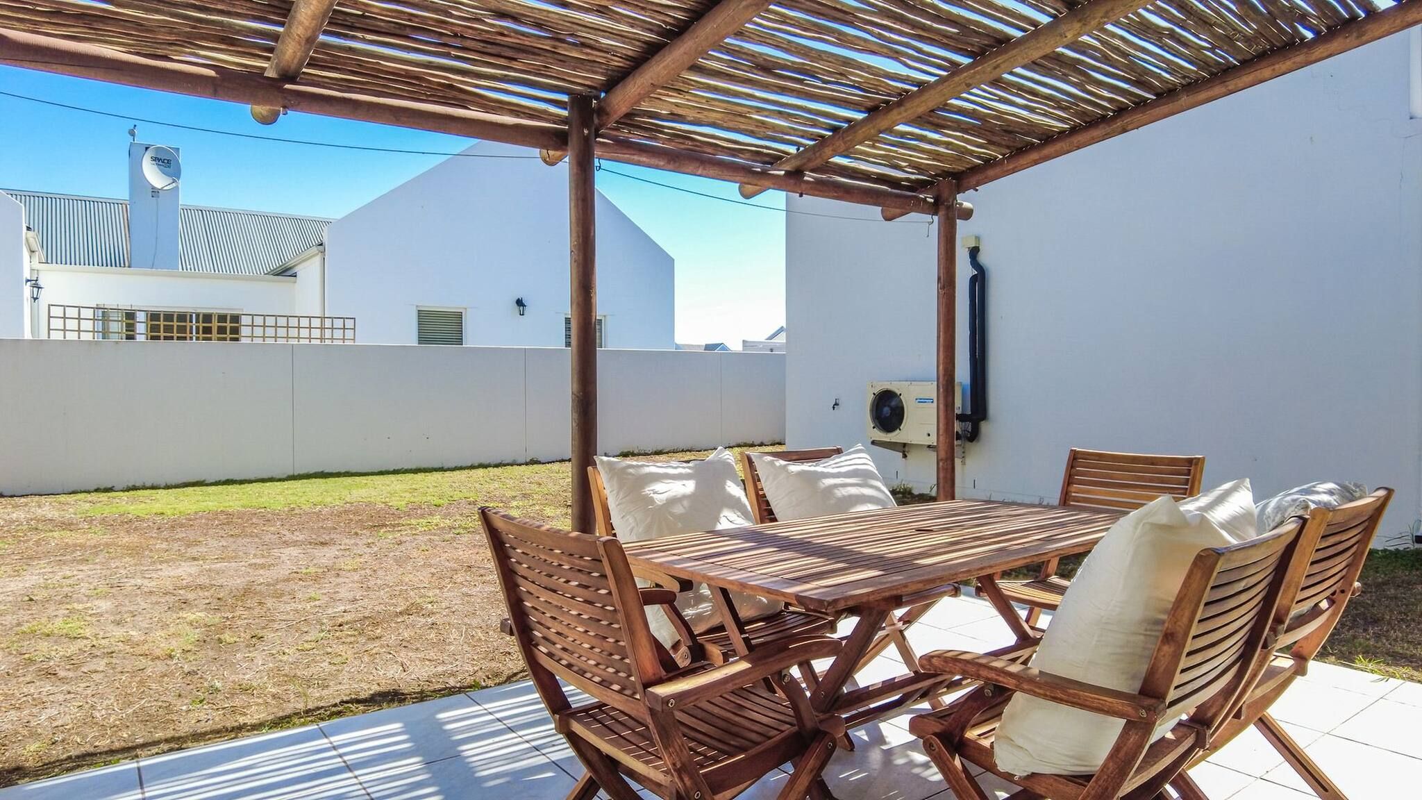 Une terrasse extérieure avec une table et des chaises en bois, une pergola au-dessus et un mur blanc en arrière-plan.