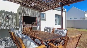 Eine Terrasse im Freien mit einem Holztisch und Stühlen, einem an der Wand befestigten Fernseher und einer Pergola darüber.