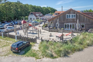 A beachside wooden building with outdoor seating, a sandy area with a slide, and a parking lot with cars. - Modernly furnished chalet in the heart of Texel (Den Burg)