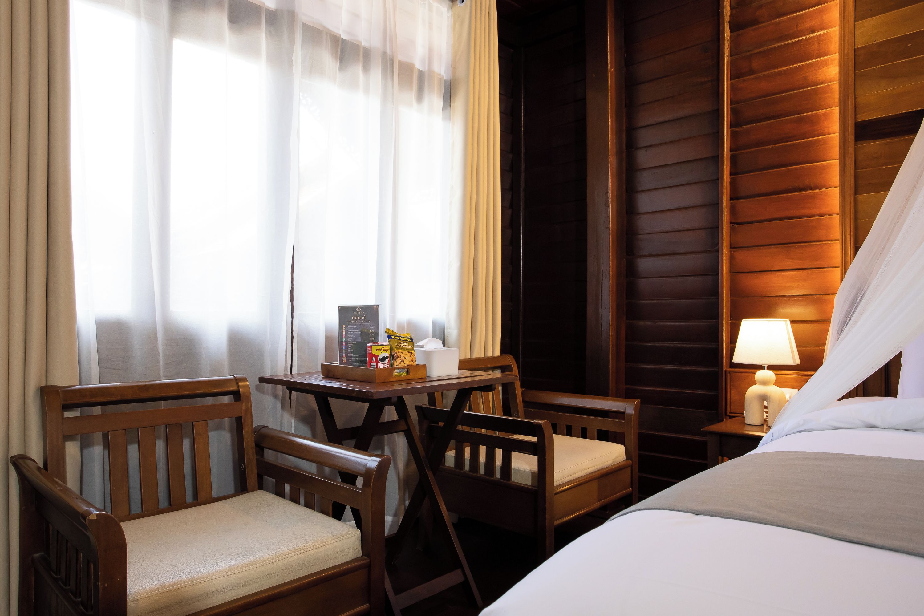 Habitación de hotel con una cama, dos sillas de madera, una mesita con aperitivos, una lámpara y una ventana con cortinas translúcidas.