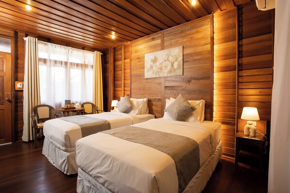 A hotel room with two beds, wooden walls, a painting, a window with curtains, a small table with a lamp, and a chair.