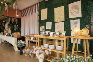 Un área de recepción con una pared verde decorada con motivos florales, un mostrador de recepción con un cartel de 'Welcome' y una mesa de buffet con diversas opciones de comida y etiquetas.