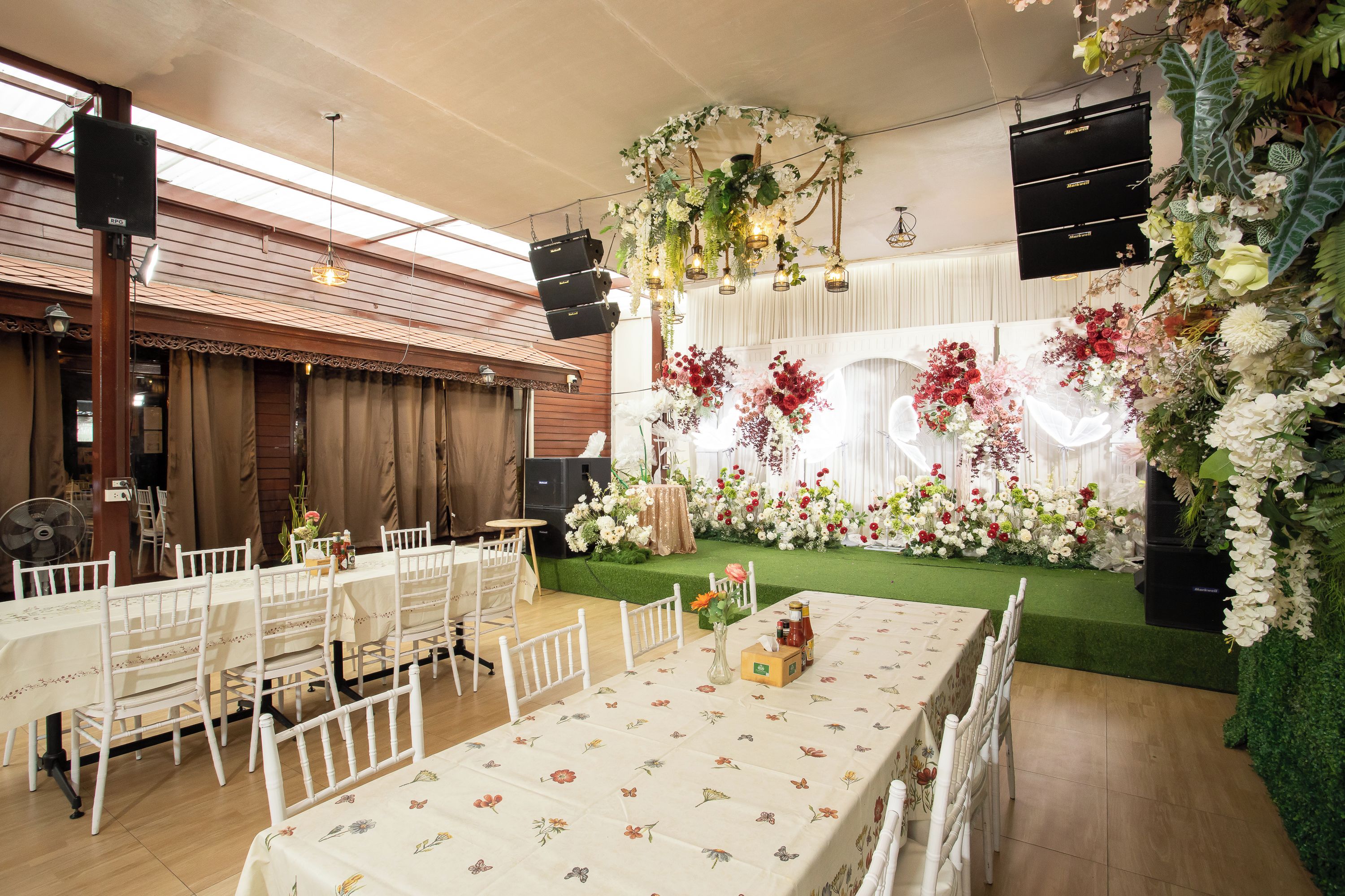 Ein Festsaal mit gedeckten Tischen für eine formelle Veranstaltung, einer Bühne mit Blumendekoration und einer Decke mit freiliegenden Balken und Beleuchtungselementen.
