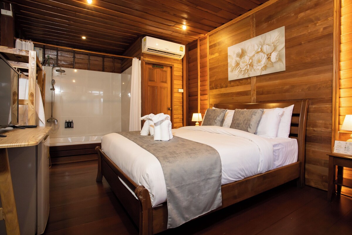 Habitación de hotel con techo de madera, cama con ropa de cama blanca, mesita de noche con lámpara y zona de ducha.