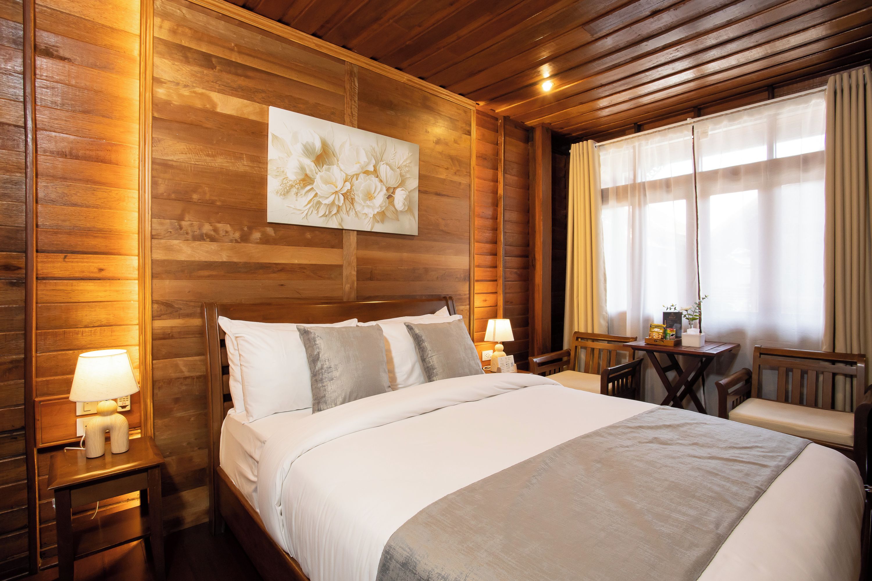 A wooden-paneled room with a bed, bedside lamps, a table with a chair, and a window with curtains.