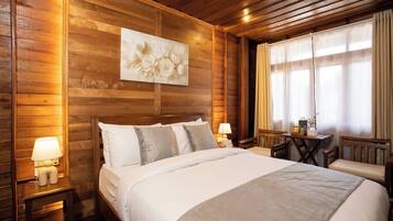A wooden-paneled room with a bed, bedside lamps, a table with a chair, and a window with curtains.