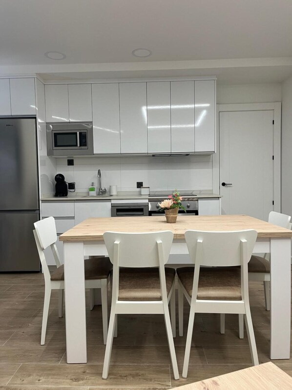 A modern kitchen with white cabinets, a stainless steel refrigerator, a microwave, and a dining area with a wooden table and white chairs.