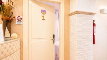 Ingresso di una camera d'albergo con una porta bianca, un cartello 'no smoking', un estintore e un vaso decorativo con rami.