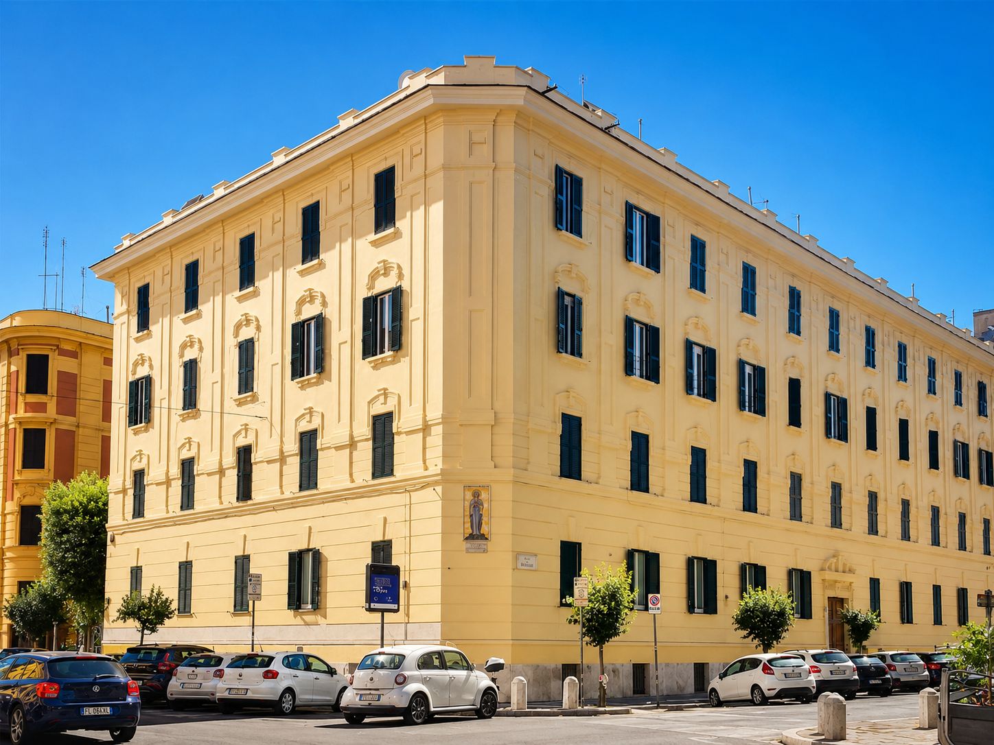 Un edificio giallo a più piani con un ingresso ad angolo, auto parcheggiate e un cielo azzurro intenso.